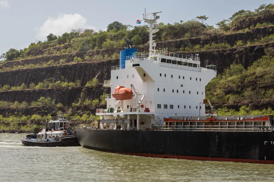 Cargo ship with a tugboat passing terraced rock walls of the Culebra Cut